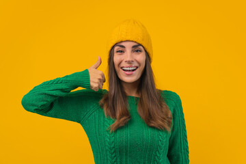 Close up portrait of happy smiling attractive young woman in green sweater and knitted yellow beanie hat showing thumbs up with excited face expression