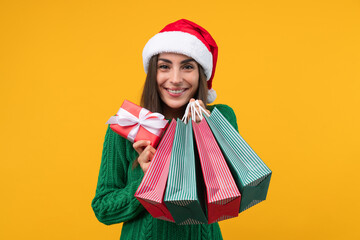 Studio portrait of attractive smiling young woman in Santa's hat posing with a gift box and a bunch of shopping bags in hands