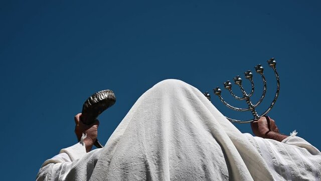 Rear view of adult Jewish man holding two Jewish symbols in his hands, a menorah (R) and a shofar (ram's horn) (L) prying for the coming of the messiah outdoors under clear blue sky.