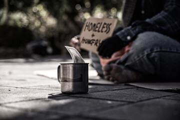 metal cup with banknotes and a homeless beggar sitting beside.
