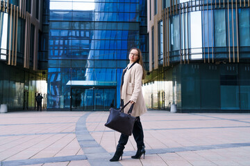 Fototapeta na wymiar Successful happy business woman in glasses, Caucasian ethnicity in stylish business clothes with a bag in her hand walks against the background of an office building on a sunny windy day