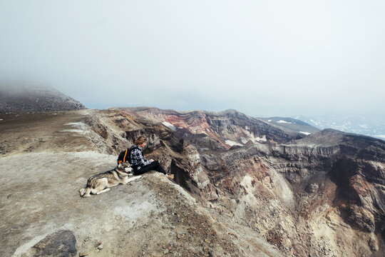 Woman with her dog sitting on the edge of active volcano crater. Beautiful view on mountain valley