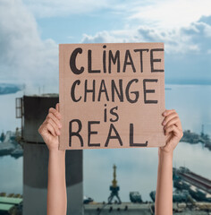Protestor holding placard with text Climate Change Is Real  and blurred view of industrial factory on background