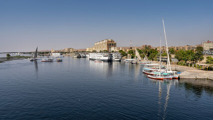 Boats and feluccas float on the smooth blue water of the Nile. There are ships at the shore. Reflection. City houses are visible in the distance. Blue sky. Egypt. Aswan.
