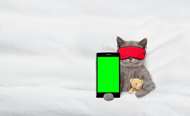 Kitten wearing mask for sleep hugs toy bear on a bed at home and shows empty screen of a...