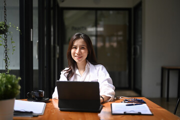 Fototapeta premium Positive female doctor in white medical uniform sitting at her workplace and smiling to camera.