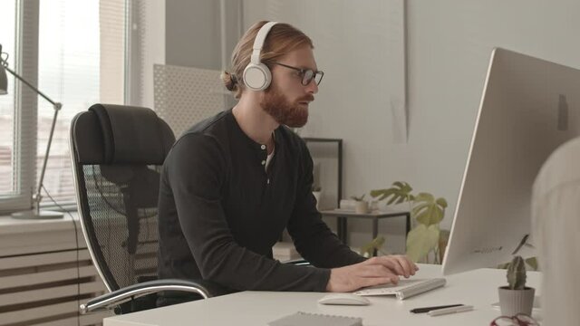 Slowmo shot of young male programmer in headphones and eyeglasses working on PC and talking to colleague sitting at table in modern office