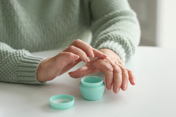 Elderly woman applying cosmetic cream onto her hands at table