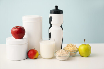 Glass of protein shake, bowls with powder, apples, jars and bottle on white table against color background