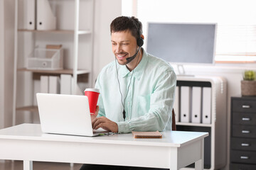 Consultant of call center with headset with cup of coffee working at table in office