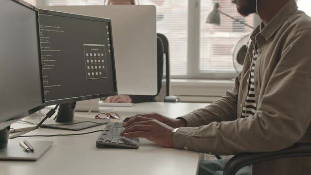 Cropped slowmo shot of African-American male programmer in casualwear and earphones writing program code on PC at modern office