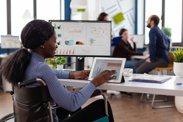 Handicapped african american businesswoman in wheelchair analyzing financial graph on tablet working at company strategy in startup office. Paralyzed manager planning business partnership