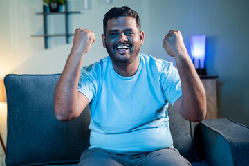 Exited Fat man watching sports on TV at night while sitting at home - Concept of cricket game supporter, watching live streaming match and weekend leisure activities
