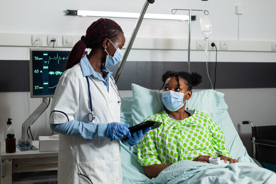 African American Practitioner Doctor With Protective Face Mask Against Covid19 Holding Tablet Computer With Medical Expertise Explaining Healthcare Treatment To Sick Patient. Medicine Service