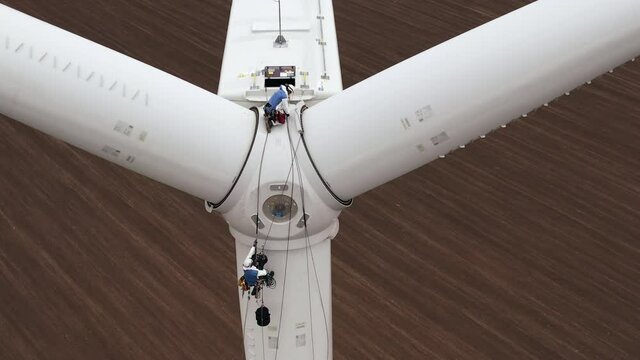 Service engineer and coworker high-altitude installer with safety belts go down to windmill propeller fastened by ropes above plowed field aerial view