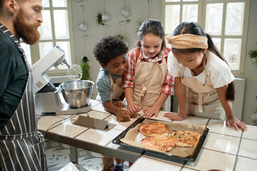 Fototapeta na wymiar Group of children in aprons looking at baked pizza on the tray and smiling, they cooking together with the chef