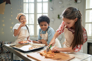 Little girl in apron cutting homemade pizza on the wooden board, children preparing pizza together in the kitchen