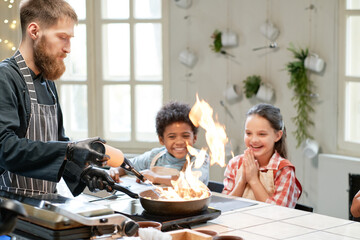 The chef cooking the food on the pan on the stove with fire and showing the masterclass to children who sitting at the table