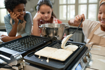 Little girl learning to bake waffles during a lesson with other children in the background