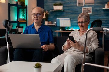 Married couple enjoying retirement with technology at home. Elder husband using laptop and old woman holding smartphone while sitting together on living room couch. Senior people with devices