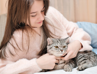 young woman with long hair lying on a bed and grey tabby cat sitting next to her. living with pet friends, relaxing together