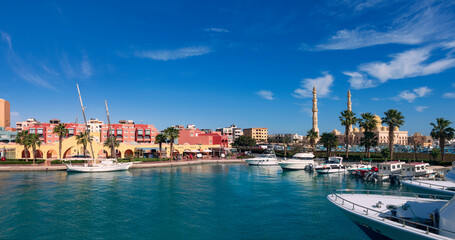 Obraz premium Panoramic view from the marina of Hurghada to the minarets of the El Mina mosque and the city's embankment with moored yachts, shops, cafes and a pedestrian zone for walking