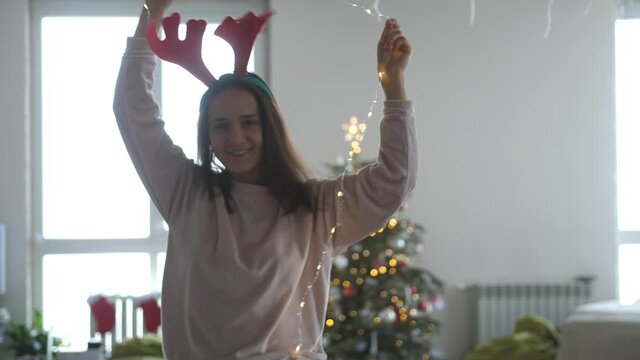 Young beautiful woman wearing reindeer antlers headband and garland dancing next to Christmas tree