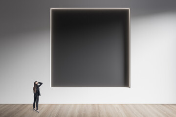 Businesswoman standing in minimalistic concrete and wooden interior with abstract clean black square frame on wall. Mock up. Exhibition, gallery and art concept.