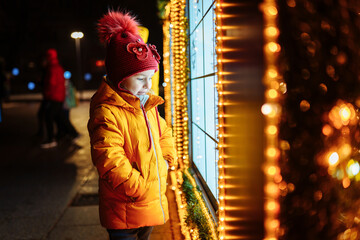 Little girl in red hat look from the street into a christmas decorated shop window on the evening before christmas