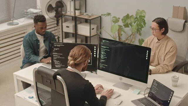 From-above slowmo shot of diverse male programmers team working on program coding sitting at one table in front of computers in modern open space office