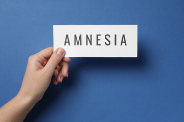 Woman holding white card with word Amnesia on blue background, top view