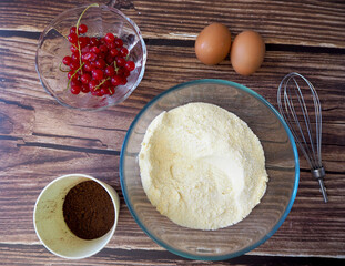 a glass bowl with corn and rice flour, sugar stands on a wooden table next to a plate with red currants, two eggs and a glass of cocoa.top view. recipe and preparation of vegan cupcake