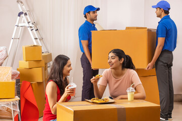 Adult girls eating pizza and drinking shake while the delivery boys moving cardboard boxes