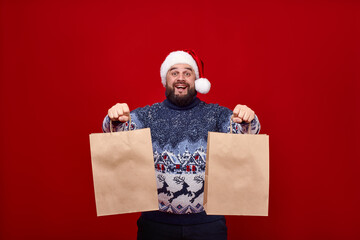 An emotional man in a Santa hat holds out two craft packages to the camera on a red background. A...