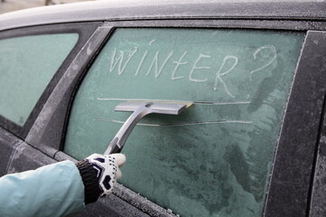 Inscription word WINTER? on snowy side car glass and woman hand with glove and scraper wiper at...