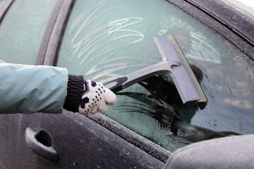 Woman's hand with warm glove  and silicone scraper cleans the car side door window with a hoarfrost...