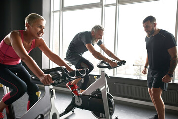 Fototapeta na wymiar Young sportive instructor watching couple cycling in gym