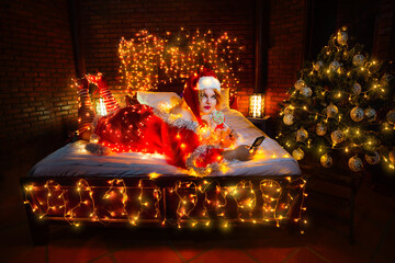 Seductive plump Santa helper holding lolly-pop lying on bed with mobile