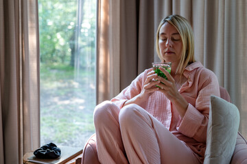 A blond woman in pink pajama sits in the pink armchair and sips mint tea.