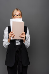 Blonde businesswoman in eyeglasses holding paper folders near face isolated on grey