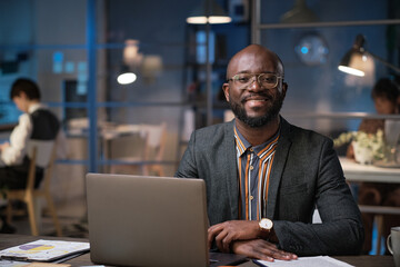 Portrait of African businessman smiling at camera while sitting at his workplace with laptop at...
