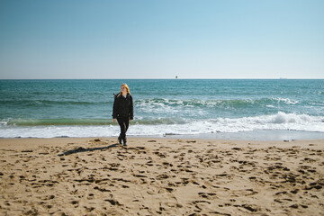 Blond girl in dark clothes with backpack walking on the winter beach