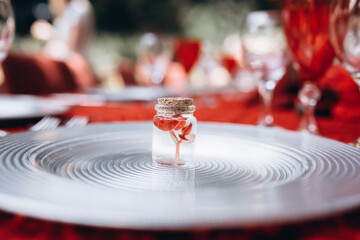 a small red flower in a transparent small jar stands on a silver plate against the background of a red tablecloth and table setting items