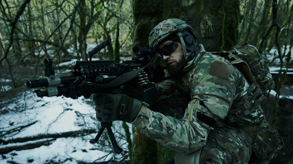 Portrait of airsoft player in professional equipment in helmet aiming at victim with gun in the forest. Soldier with weapons at war