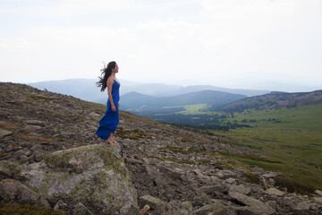 A beautiful woman of Asian appearance in a blue dress stands on the edge of a cliff among the stones in the Altai mountains in summer. concept beauty, tourism.