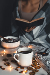 The woman is wrapped in a blanket and holds a book in her hands. cup of coffee . Winter comfort, garlands, Christmas mood. Stylish manicure,