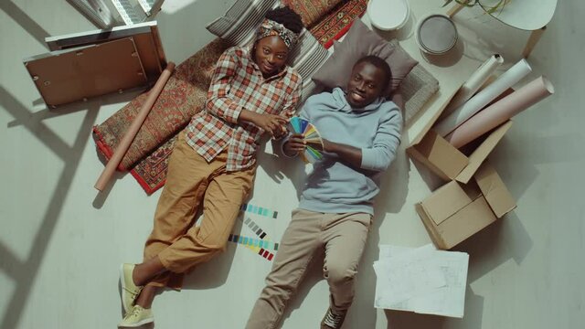 Top down shot of African American couple lying on floor with home renovation supplies around, holding color palette and looking at camera with smile