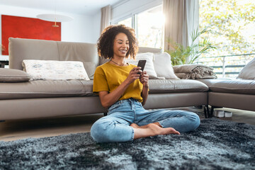 Obraz premium Happy mature woman using smartphone while relaxing sitting on the floor at home.