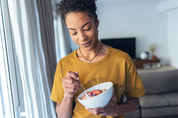 Beautiful mature woman eating cereals and fruits while standing next to the window at home.