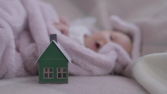 Close-up toy house on cozy bed with blurred innocent baby girl sleeping at background. Newborn Caucasian child enjoying nap indoors at home. Leisure and happiness